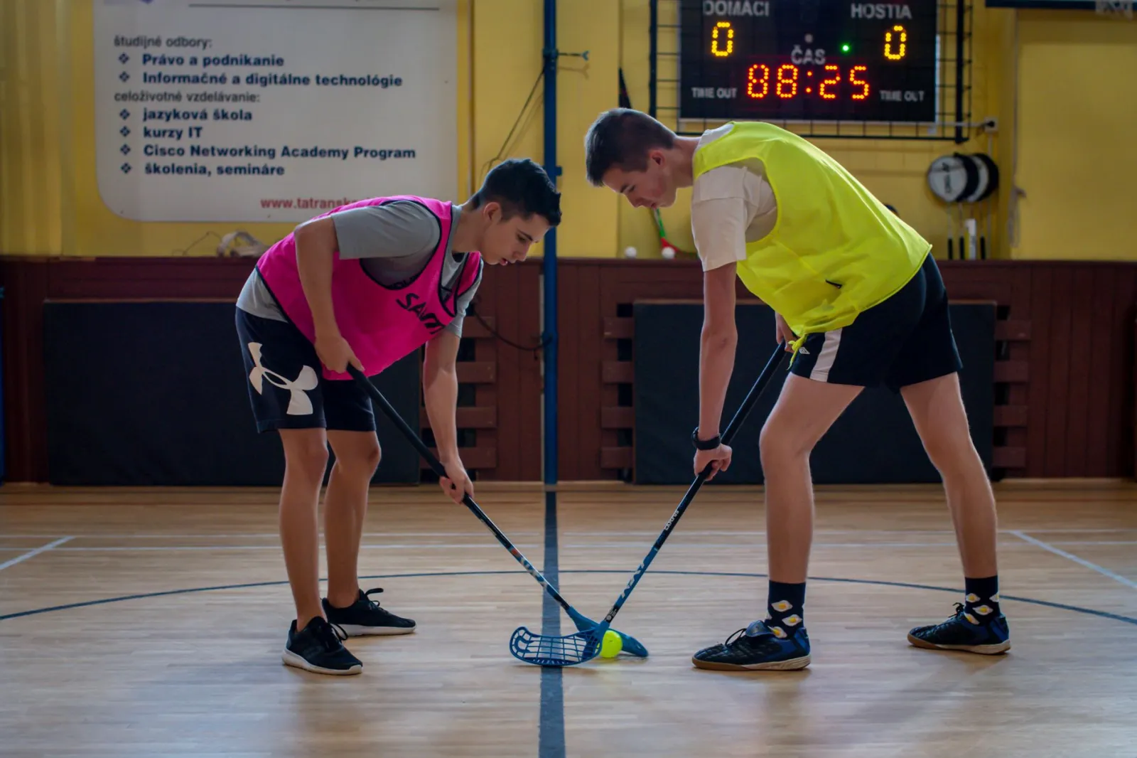 Two teenagers face off in a floorball match inside a sports gym with equipment visible in the background.